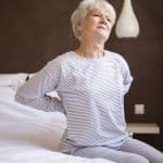 Mulher branca idosa com as mãos nas costas demonstrando dores por conta da osteoporose