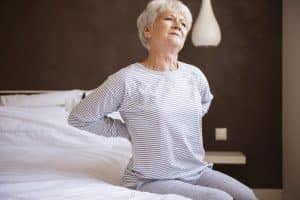 Mulher branca idosa com as mãos nas costas demonstrando dores por conta da osteoporose