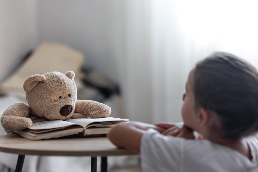 Criança observa um ursinho de pelúcia apoiado sobre um livro aberto, simbolizando o poder do brincar e da imaginação no desenvolvimento emocional e na saúde mental infantil