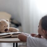 Criança observa um ursinho de pelúcia apoiado sobre um livro aberto, simbolizando o poder do brincar e da imaginação no desenvolvimento emocional e na saúde mental infantil