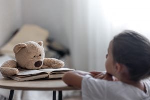 Criança observa um ursinho de pelúcia apoiado sobre um livro aberto, simbolizando o poder do brincar e da imaginação no desenvolvimento emocional e na saúde mental infantil
