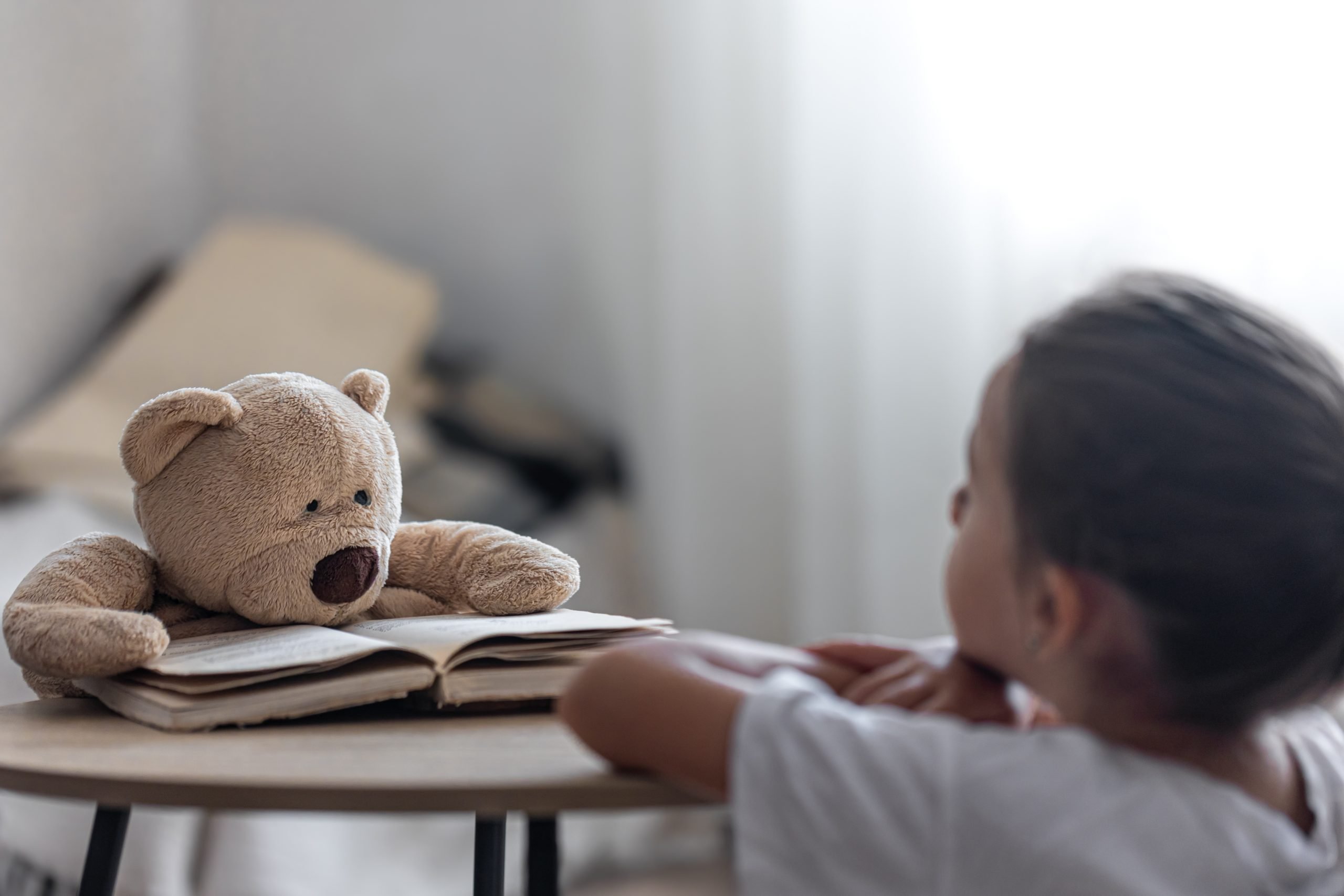 Criança observa um ursinho de pelúcia apoiado sobre um livro aberto, simbolizando o poder do brincar e da imaginação no desenvolvimento emocional e na saúde mental infantil
