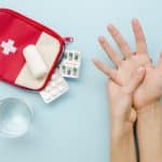 Kit de primeiros socorros com curativos, comprimidos e água ao lado de uma pessoa segurando a própria mão, ilustrando cuidados básicos em situações de emergência do dia a dia