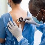Médico negro com avental cirúrgico azul e luvas brancas examina pinta nas costas de paciente branco ao investigar câncer de pele