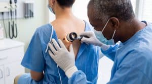 Médico negro com avental cirúrgico azul e luvas brancas examina pinta nas costas de paciente branco ao investigar câncer de pele