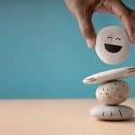 Mão equilibrando pedras empilhadas com um rosto sorridente no topo, simbolizando regulação emocional e o cuidado em organizar e lidar melhor com as emoções.