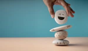 Mão equilibrando pedras empilhadas com um rosto sorridente no topo, simbolizando regulação emocional e o cuidado em organizar e lidar melhor com as emoções.