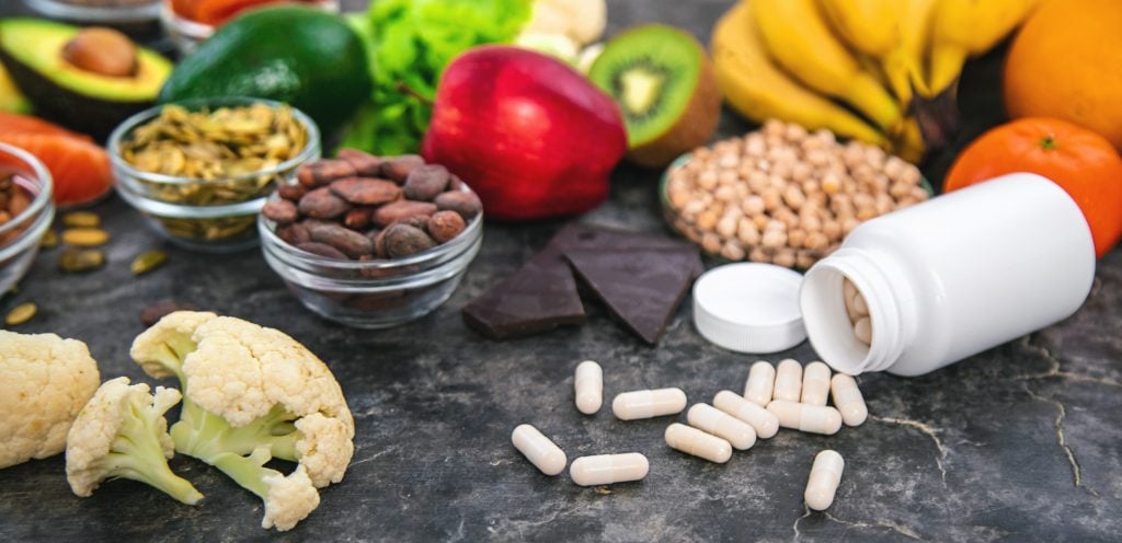 Alimentos ricos em magnésio, como abacate, banana, sementes, cacau, feijão e vegetais, ao lado de cápsulas de suplemento sobre a mesa.
