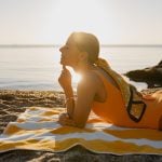 Mulher deitada na areia da praia durante o pôr do sol, aproveitando o sol para bronzeamento, destacando cuidados com a pele no verão.