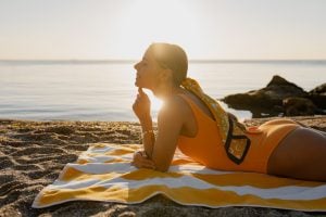 Mulher deitada na areia da praia durante o pôr do sol, aproveitando o sol para bronzeamento, destacando cuidados com a pele no verão.