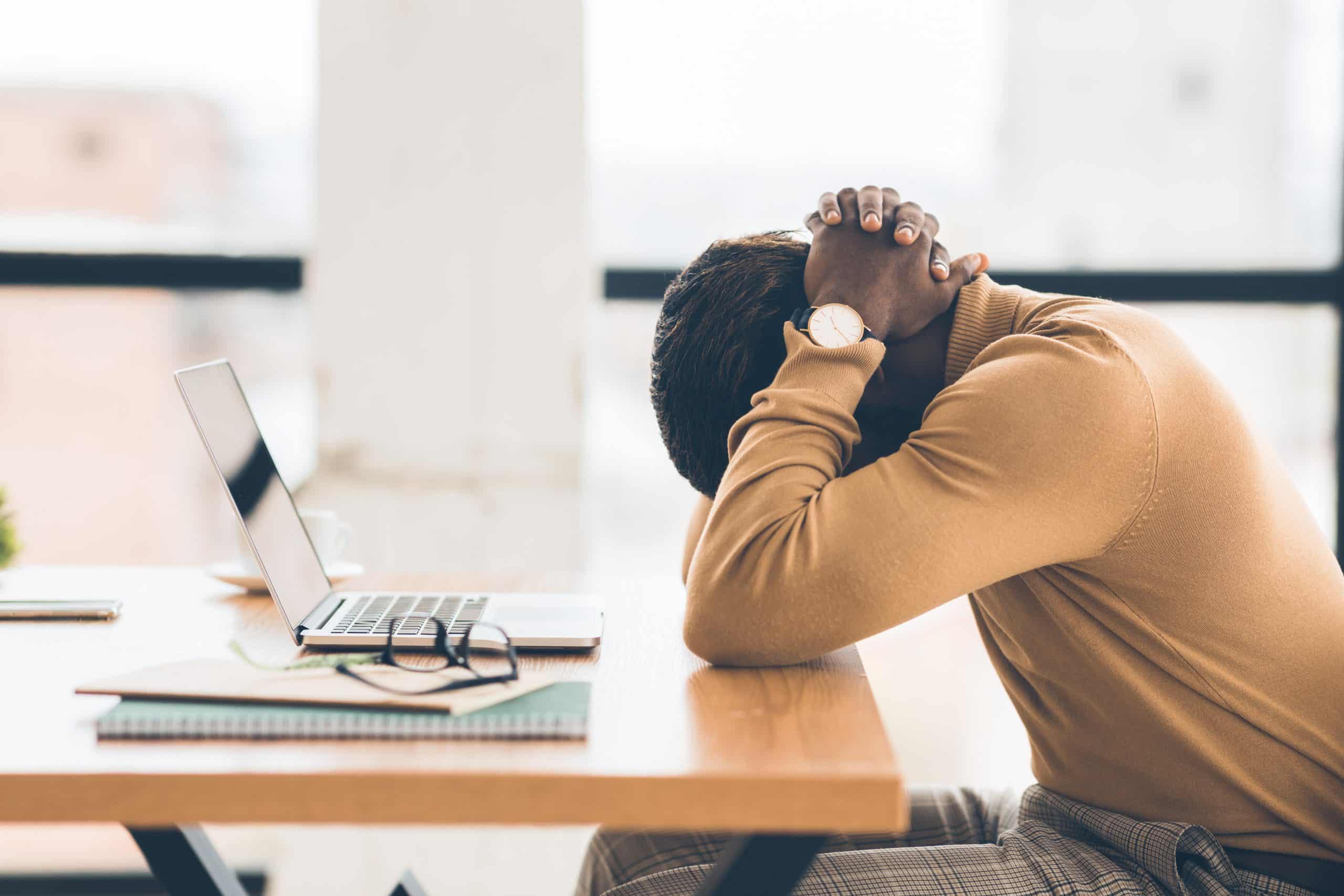 Pessoa sentada à mesa de trabalho com a cabeça apoiada nas mãos ao lado de um notebook, demonstrando cansaço e esgotamento mental, sinais comuns de burnout no dia a dia.