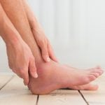 Pessoa sentada segura o tornozelo com as mãos, evidenciando inchaço na região do pé e da articulação, em ambiente interno com piso de madeira claro