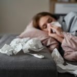 Pessoa com sintomas de gripe K deitada no sofá assoando o nariz, com termômetro e lenços usados ao lado.