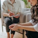 Mulher em atendimento psicológico, com expressão preocupada, enquanto profissional faz anotações, representando a importância da saúde mental durante a busca por emprego.