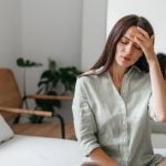 Mulher com a mão na testa e expressão de desconforto, indicando dor de cabeça, em ambiente doméstico.