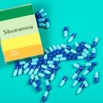 Caixa de sibutramina com indicação de 10 mg e cápsulas azuis espalhadas sobre fundo verde, representando medicamento usado no controle de peso.