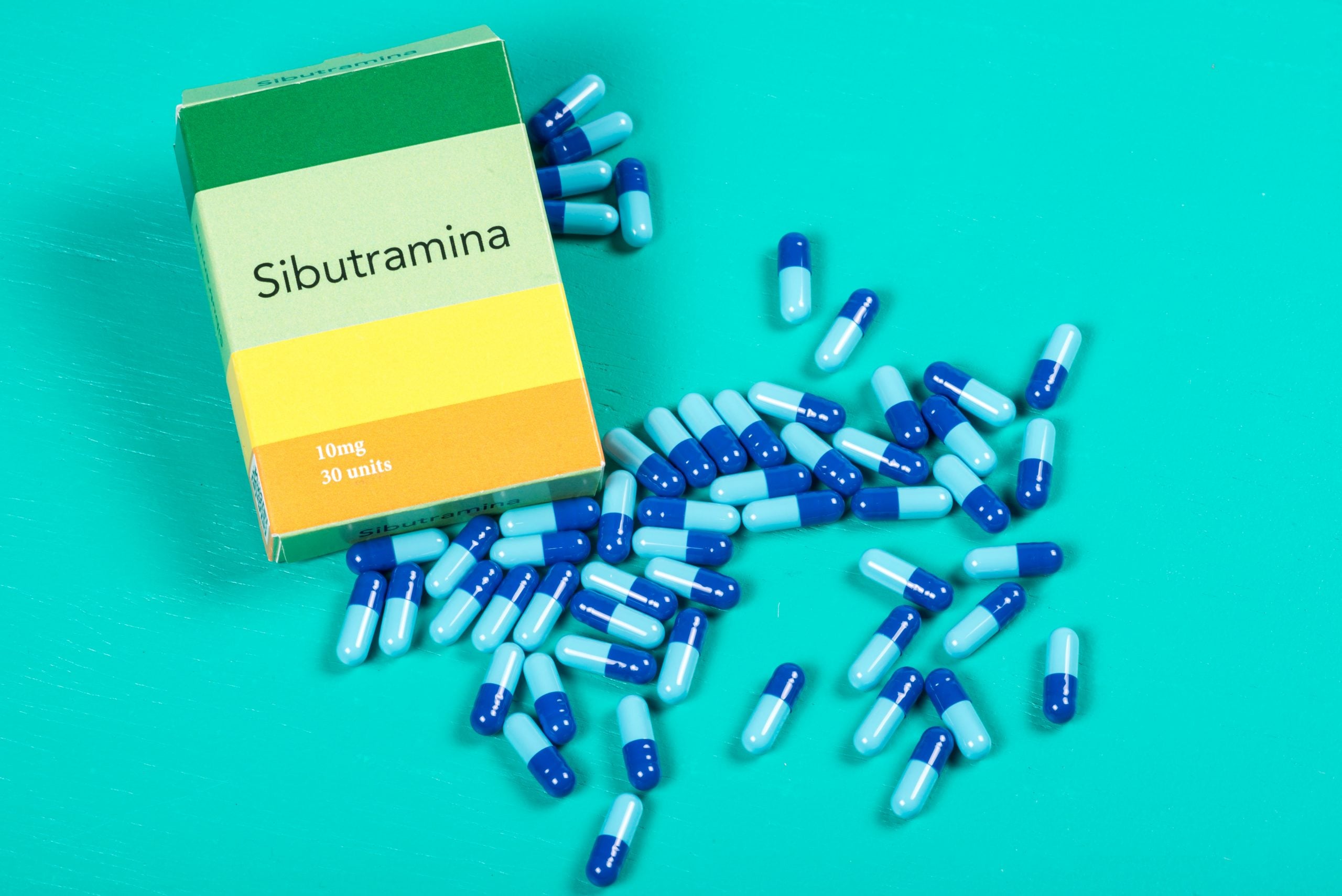 Caixa de sibutramina com indicação de 10 mg e cápsulas azuis espalhadas sobre fundo verde, representando medicamento usado no controle de peso.