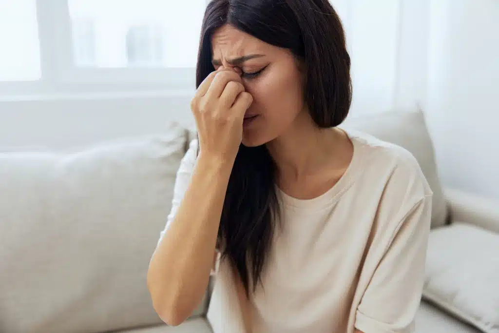 Mulher com expressão de dor pressionando a região do nariz e dos olhos, ilustrando sintomas comuns da Sinusite, como pressão facial e desconforto nasal.