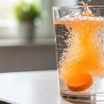 Comprimido efervescente de Vitamina C dissolvendo-se em um copo de água, liberando bolhas, ilustrando o uso do suplemento durante períodos de resfriado, gripe e baixa imunidade.