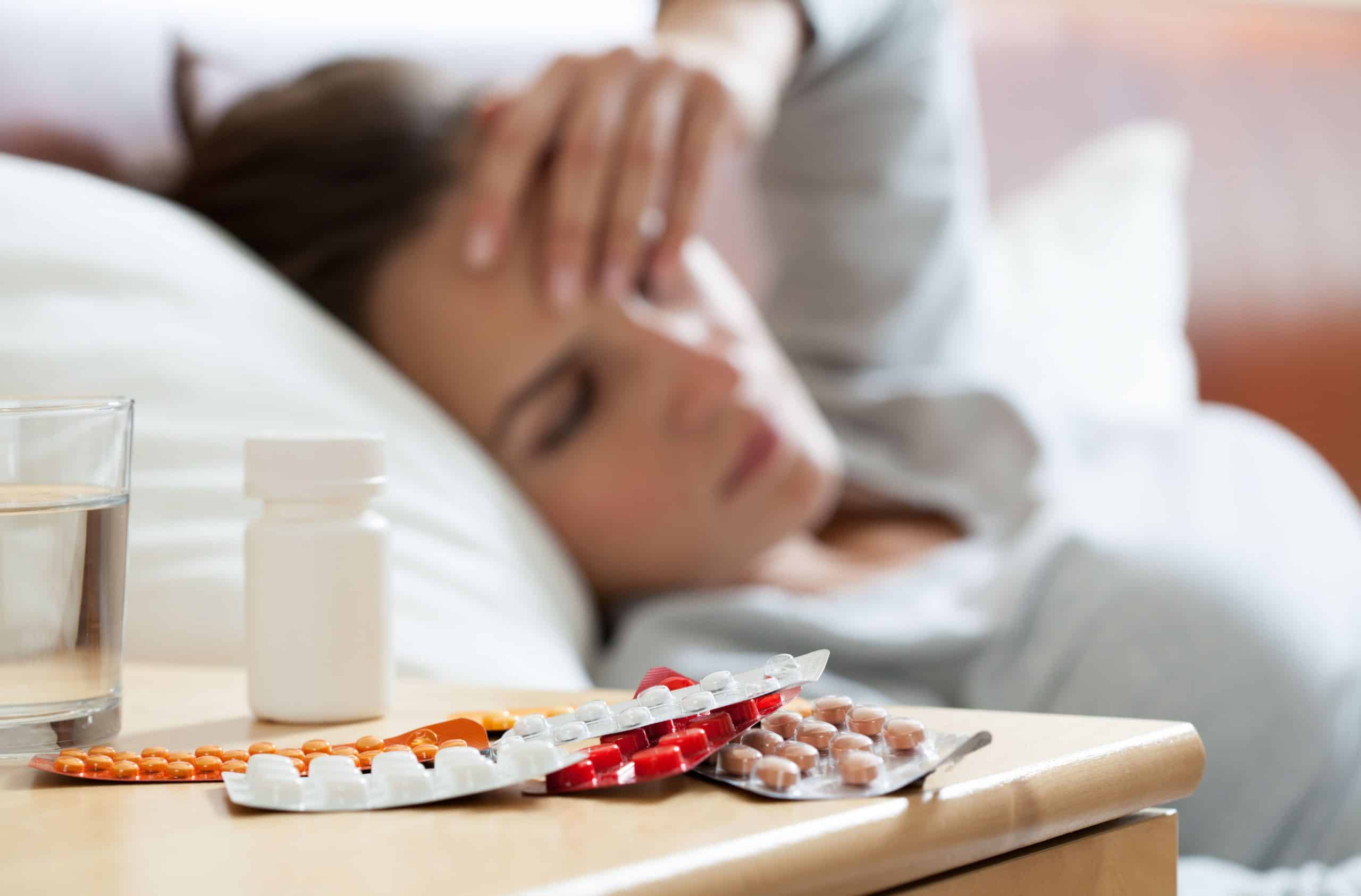 Pessoa deitada com sintomas de gripe, segurando a testa enquanto medicamentos e um copo de água estão sobre a mesa de cabeceira, ilustrando o uso de remédios para aliviar os sintomas da gripe.